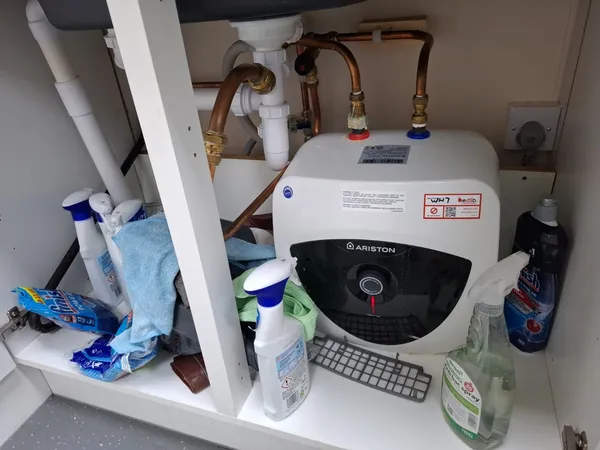 install — white Ariston Andris Lux 10-litre under-sink point-of-use electric water heater fitted under a UK kitchen sink, with twin 15 mm copper feeds, red and blue colour-coded isolation valves and an edSb commissioning sticker.