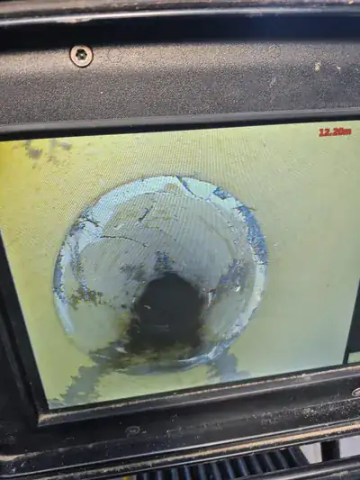 CCTV drain survey monitor — view down a 100mm clay underground drain pipe at 12.20 metres showing a cracked section and a displaced joint with visible debris build-up before patch liner repair.