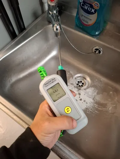 TMV calibration — gloved hand holding an ETI TempTest Blue smart thermometer with a coiled stainless probe inserted into the running stream of a stainless steel sink mixer tap during a thermostatic mixing valve temperature verification.
