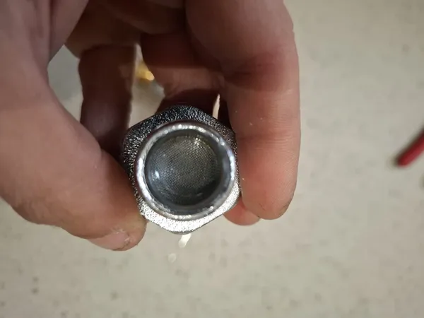 TMV service — engineer's hand holding a chrome-plated TMV inlet strainer body showing a fine stainless steel filter mesh after being cleaned during a scheduled thermostatic mixing valve service.